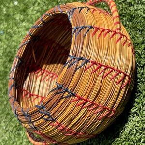 May include: Handmade woven basket with a rounded shape and a handle. The basket is made of natural-colored reeds, with dark brown accents and red and navy blue stitching details. The basket is sitting on a green surface.