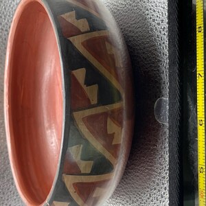 May include: A hand-painted wooden bowl with a reddish-orange interior and a black exterior band. The exterior features a repeating geometric pattern in red, tan, and green. The bowl is approximately 7 inches in diameter.