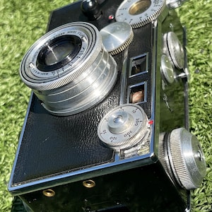 May include: A vintage black camera with silver-colored dials and a lens. The camera has a rectangular body with rounded edges and is resting on a green surface. The lens is labeled "72.5 50MM ARGUS COATED CINTAR MADE IN USA".