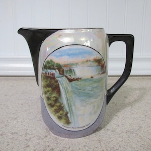 May include: A white ceramic pitcher with a black handle and a colorful image of Niagara Falls. The pitcher has a pearlescent finish and the text "Prospect - Palm Niagara Falls" is printed on the bottom.
