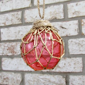 May include: A pink glass float encased in a natural twine net, hanging from a twine cord. The float is a traditional nautical decoration.