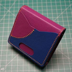 May include: A bi-fold wallet in a vibrant color scheme. The top section is a deep magenta, contrasted by a navy blue lower section. The two sections are stitched together with gold thread. A small cutout is visible on the blue section.