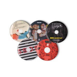 Puede incluir: Cinco CD de la banda 5 Seconds of Summer. Los CD tienen varios diseños, incluyendo fotos de la banda, texto y gráficos. Los colores incluyen rojo, negro, blanco y beige. Los CD están dispuestos en un patrón circular.