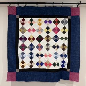 May include: Handmade quilt with a white center featuring a diamond pattern of various colors, including pink, blue, yellow, and brown. The quilt is framed by a black border, a dark blue border, and pink corner accents. It is hanging on a rod.
