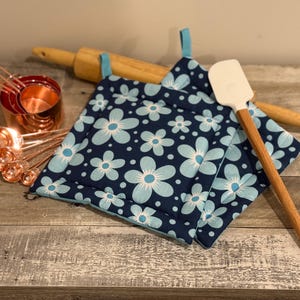 May include: A collection of kitchen tools and accessories, including copper measuring cups and spoons, a wooden rolling pin, a white spatula with a wooden handle, and two navy blue pot holders with a light blue floral pattern.