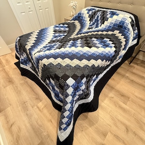 May include: A handmade quilt with a geometric pattern in shades of blue, black, and white. The quilt is draped over a bed, showcasing its intricate design and craftsmanship. The quilt's border is black.