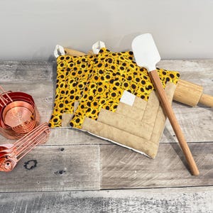 May include: A collection of kitchen tools and accessories, including copper measuring cups and spoons, a white spatula with a wooden handle, a wooden rolling pin, and two sunflower-patterned oven mitts. The items are arranged on a rustic wooden surface.