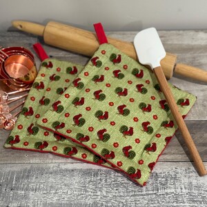 May include: Two green and red rooster-themed pot holders with red trim, a wooden rolling pin, a white spatula, and copper measuring cups and spoons. The pot holders have a red loop for hanging.