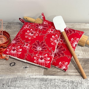 May include: A set of copper measuring cups and spoons, two red floral print pot holders, a wooden rolling pin, and a white spatula with a wooden handle. The pot holders have a white floral design.