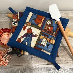 May include: A collection of fall-themed kitchen items. Includes a blue-trimmed pot holder with a pumpkin scarecrow design, copper measuring cups and spoons, and a white spatula with a wooden handle. A rolling pin is also visible.