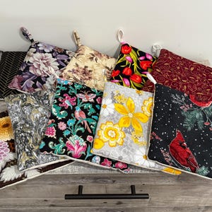 May include: A collection of colorful, square pot holders with hanging loops. Designs include floral patterns, a cardinal bird, and geometric shapes. The pot holders are displayed on a wooden surface.