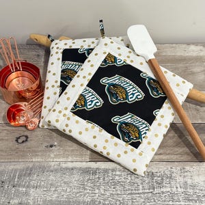 May include: Two square pot holders with a black center featuring the Jacksonville Jaguars logo, surrounded by a white fabric with gold polka dots. A wooden rolling pin and a white spatula with a wooden handle are also present. Copper measuring cups and spoons are on the left.