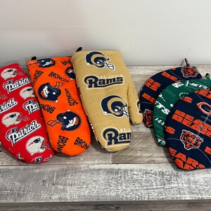 May include: A collection of oven mitts and pot holders featuring NFL team logos. The mitts and pot holders are in red, orange, tan, green, and navy blue. The logos include the Patriots, Broncos, Rams, and Bears.