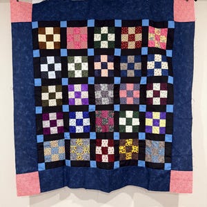 May include: A handmade quilt with a patchwork design. The quilt features a dark blue border and a central section composed of numerous squares in various colors and patterns, including floral and geometric designs. Pink corner accents add a contrasting touch.