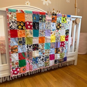 May include: A colorful patchwork quilt draped over a white crib. The quilt is made of various fabric squares in bright colors and patterns, including polka dots, cartoon characters, and floral designs. The quilt's edge is trimmed with a pink border and fringe.