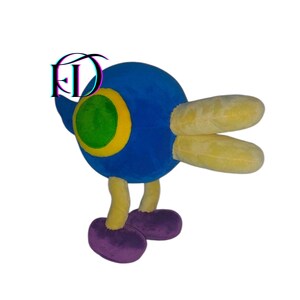 May include: A plush toy in the shape of a cartoon character. The toy has a blue body, yellow wings, and yellow legs. The large eye is green with a yellow outline, and the feet are purple.