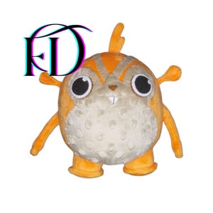 May include: A plush toy in the shape of a round, orange creature with large black eyes, small arms, and feet. The toy has a beige belly with small, textured bumps. The letters "FD" are in the upper left corner.