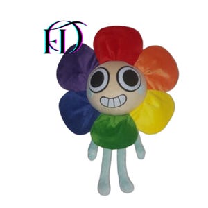 Op de afbeelding: Een pluche speelgoed in de vorm van een bloem met een lachend gezicht. De bloemblaadjes zijn rood, oranje, geel, groen, paars en blauw. Het gezicht is beige met grote zwarte ogen en een brede grijns. Het heeft lichtblauwe armen en benen.