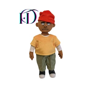 May include: A handmade doll with a serious expression, wearing a red knit cap, yellow t-shirt, white long-sleeve shirt, green corduroy pants, and black and white sneakers. The doll has brown skin and is standing against a white background.