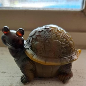 Puede incluir: Figura decorativa de tortuga con un detallado diseño de girasoles en su caparazón. La tortuga es principalmente verde oscuro con detalles amarillos y cabeza marrón. La figura es de material brillante.