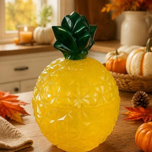 May include: A yellow glass container shaped like a pineapple with a faceted, textured surface and a removable dark green glass lid featuring pointed leaves. The object has a rounded body and a distinct geometric pattern across its exterior surface.