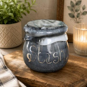 May include: A round, dark gray and white marbled ceramic jar with a matching lid. The word Stash is written in silver cursive lettering on the front of the jar. The lid features a swirling pattern of gray, white, and black tones with a glossy finish.