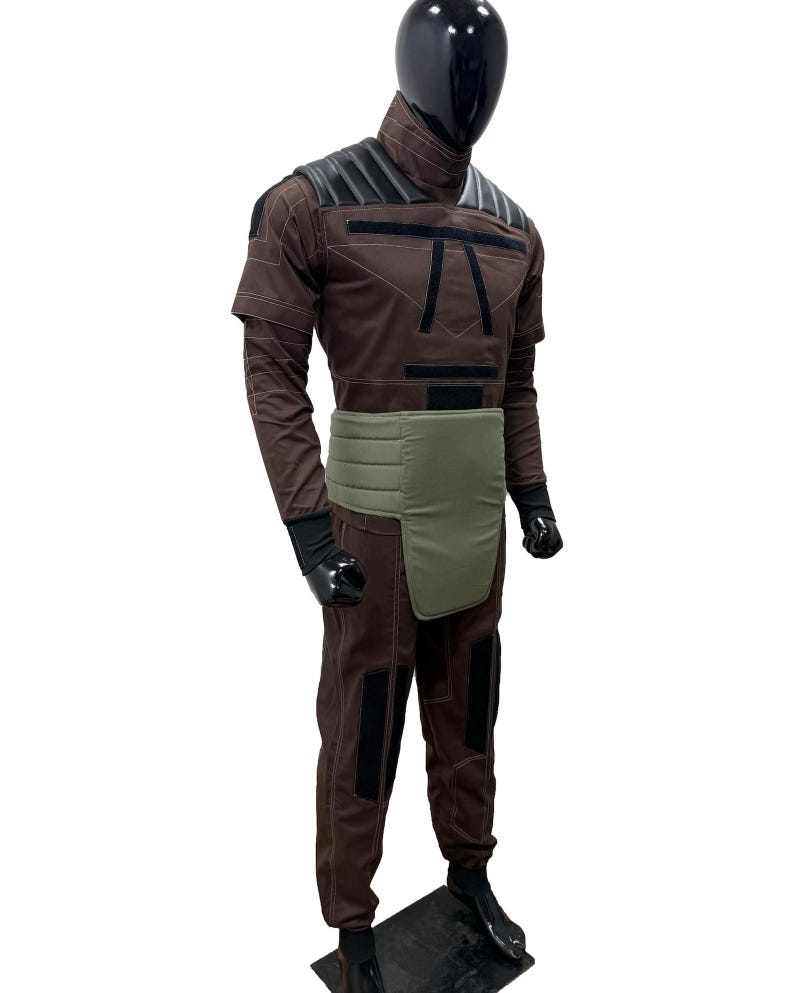May include: A brown jumpsuit with black accents, a green waist wrap, and black gloves and boots. The costume has a high collar and shoulder pads. The mannequin has a black head.