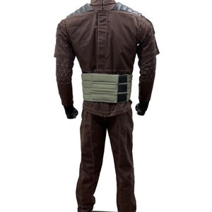 May include: A brown jumpsuit with a light green belt and black gloves. The jumpsuit has a high collar and padded shoulder details. The sleeves have a geometric pattern on the forearms. The mannequin is standing on a black surface.