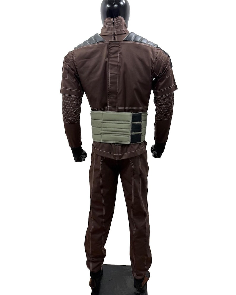 May include: A brown jumpsuit with a light green belt and black gloves. The jumpsuit has a high collar and padded shoulder details. The sleeves have a geometric pattern on the forearms. The mannequin is standing on a black surface.