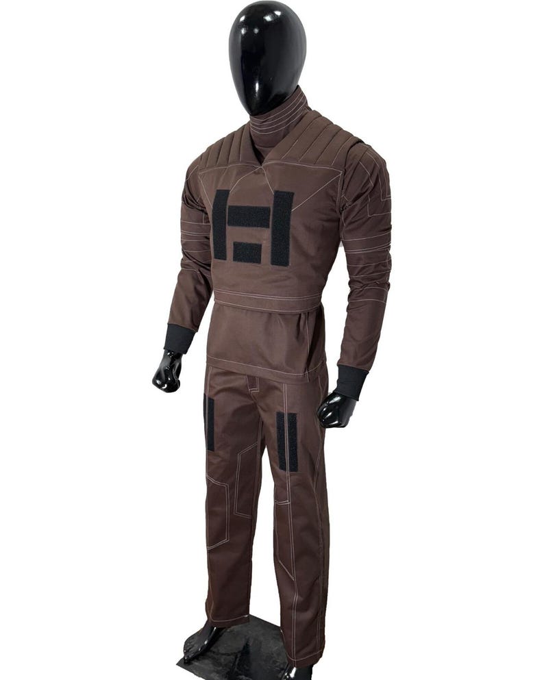 May include: Brown jumpsuit with a high collar and long sleeves. The suit features a padded chest and upper arm design, with black accents and white stitching. The pants have black patches on the knees. The mannequin is wearing black gloves.