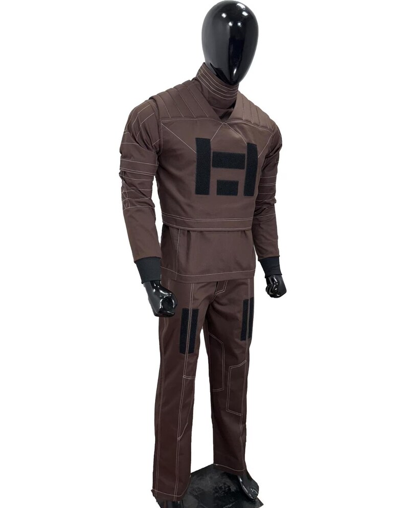 May include: Brown jumpsuit with black accents and white stitching. The outfit features a high collar, long sleeves, and a large black "H" emblem on the chest. The pants have black patches and the mannequin wears black gloves.