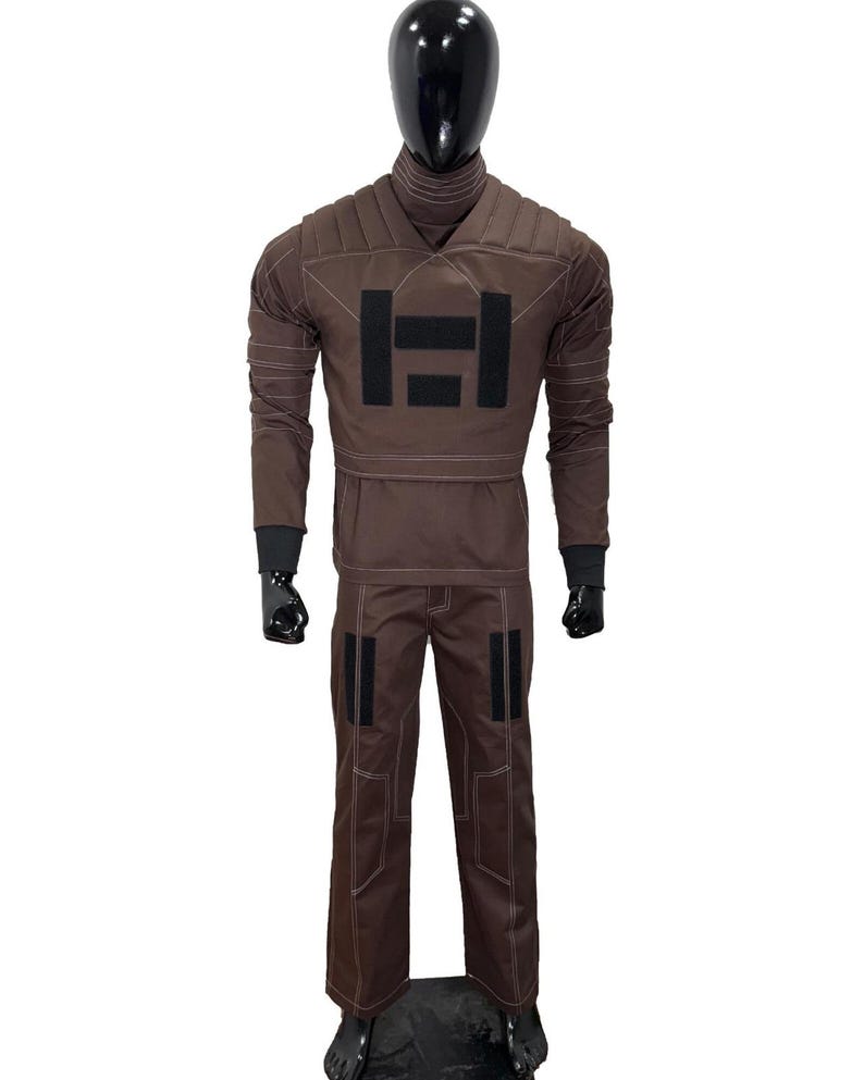 May include: A brown costume displayed on a black mannequin. The outfit includes a long-sleeved top with a black emblem and matching pants. The costume has black accents and white stitching.