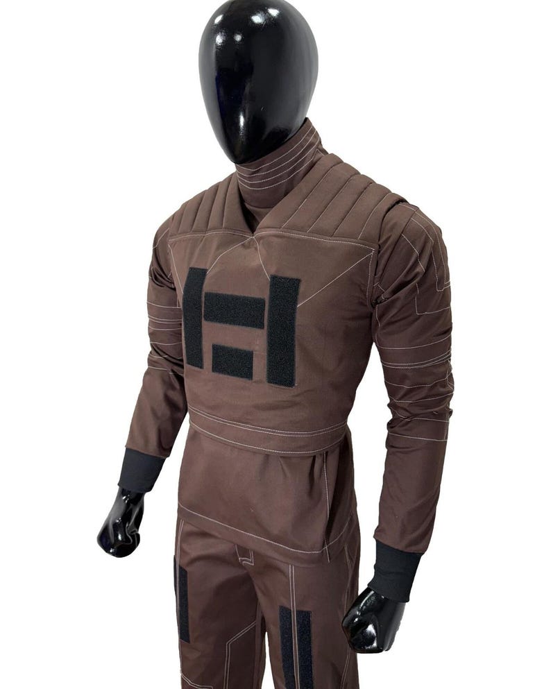 May include: Brown jumpsuit with black accents, featuring a quilted upper section and a high collar. The suit has black patches and cuffs, with white stitching details. The mannequin is wearing black gloves and a black head.