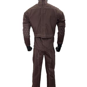 May include: A brown jumpsuit with white stitching and geometric patterns on the sleeves. The suit is displayed on a mannequin, showcasing its design and construction. The jumpsuit has a high collar and a zipper closure.
