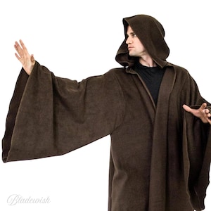 May include: A brown hooded robe with wide sleeves. The robe is made of a soft, textured fabric and has a deep hood. The person is wearing a black shirt underneath. The robe is a costume piece.