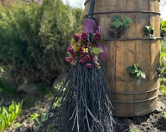 May include: A decorative black broom with a bundle of dark red flowers and purple ribbon. The broom is leaning against a weathered wooden barrel with metal bands. The scene is set outdoors with greenery in the background.
