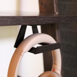 May include: A black metal headphone stand mounted under a dark wooden shelf. The stand holds a pair of beige headphones. The headphones have a curved design and are resting in the stand, ready for use.