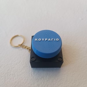 Puede incluir: Un llavero con una base cuadrada negra y una parte superior circular azul. La superficie azul tiene un acabado texturizado y muestra el texto en relieve blanco "KOYPAΓIO". Una anilla y cadena de metal dorado están unidas al lado de la base.