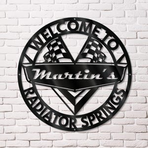 May include: Black metal sign with the words "WELCOME TO RADIATOR SPRINGS" in a circular design. The sign also features the name "Martin's" and two checkered flags. The sign is mounted on a white brick wall.