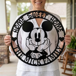 May include: Black metal circular sign featuring Mickey Mouse with the text "HOME SWEET HOME THE MICK FAMILY". The sign has two heart cutouts and is held by a person.