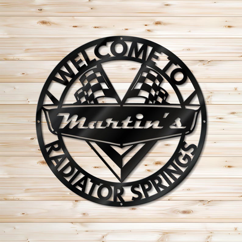 May include: Black metal sign with the words "WELCOME TO RADIATOR SPRINGS" in a circular design. The sign includes two checkered flags and the name "Martin's" in a banner. The sign is mounted on a light wood background.