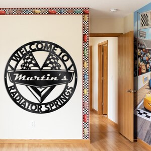 May include: A black metal wall sign that reads "WELCOME TO Martin's RADIATOR SPRINGS" with checkered flags. The sign is circular and mounted on a white wall. The room has a race car theme with decorative borders and wall art.