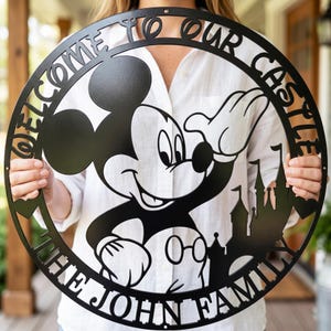 May include: Black metal circular sign featuring Mickey Mouse waving, a castle silhouette, and the text "WELCOME TO OUR CASTLE THE JOHN FAMILY". The sign is held by a person wearing a white shirt.