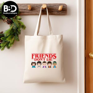 May include: A cream-colored tote bag with the text "FRIENDS DON'T LIE" in red, with cartoon character illustrations below. The bag has long handles and is hanging on a wooden rack.