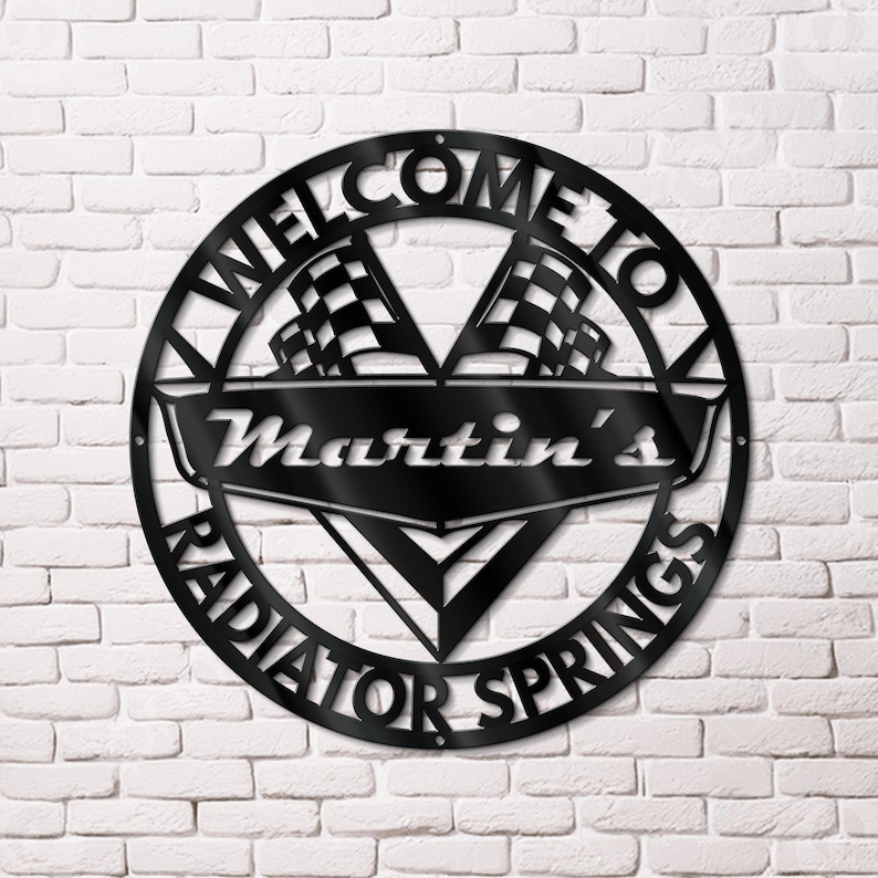 May include: Black metal sign with the words "WELCOME TO RADIATOR SPRINGS" in a circular design. The sign also features the name "Martin's" and two checkered flags. The sign is mounted on a white brick wall.