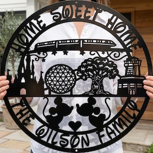 Könnte beinhalten: Schwarze Metall-Wandkunst mit den Worten "Home Sweet Home" und "The Wilson Family". Das kreisförmige Design zeigt Silhouetten eines Schlosses, eines Zuges, einer geometrischen Kugel, eines Baumes, eines Leuchtturms sowie Mickey und Minnie Mouse.