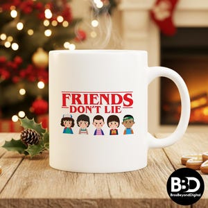 May include: White ceramic mug with the text "FRIENDS DON'T LIE" in red, above cartoon illustrations of five characters. The mug is on a wooden surface, with a Christmas tree and fireplace in the background.