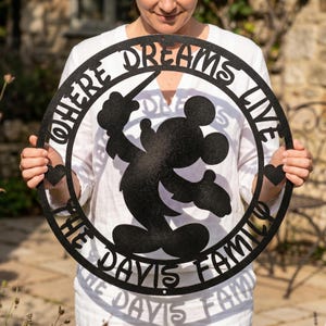 May include: Black metal sign featuring Mickey Mouse with the text "WHERE DREAMS LIVE THE DAVIS FAMILY". The sign is circular with a heart on each side. The design is cut out, creating a silhouette.