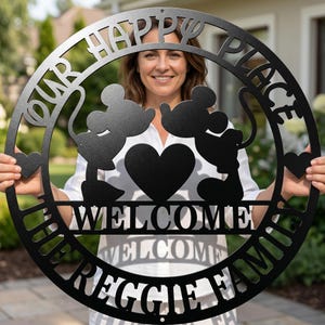May include: Black metal sign with the words "OUR HAPPY PLACE" and "WELCOME REGGIE FAMILY" cut out. Features silhouettes of Mickey and Minnie Mouse, hearts, and a circular design. A decorative item for home decor.