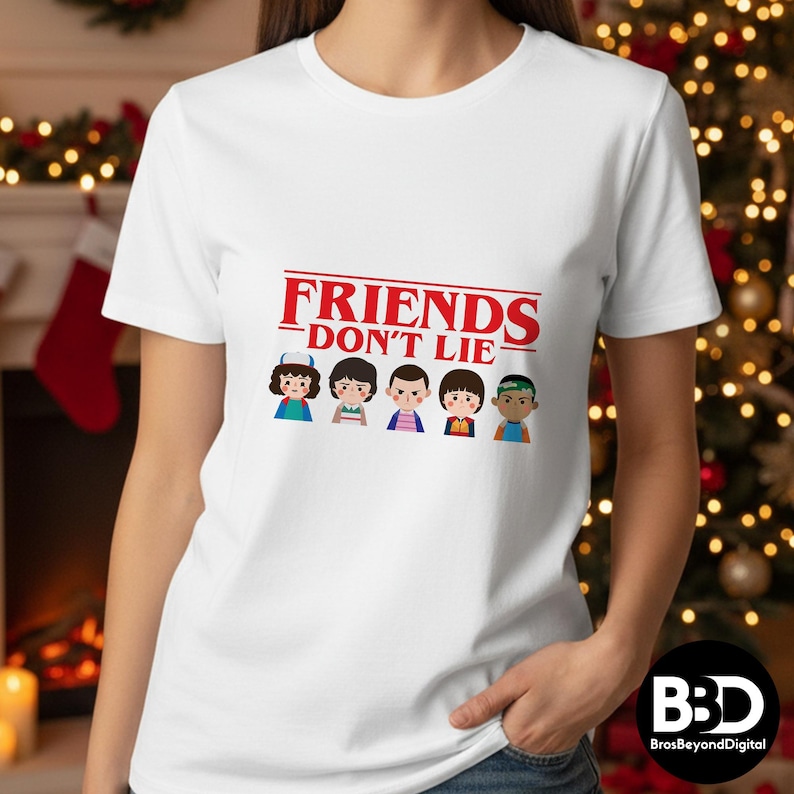 May include: White t-shirt featuring the text "FRIENDS DON'T LIE" in red, above a graphic of five cartoon characters. The characters are in various outfits and hairstyles. The shirt is a crew neck.
